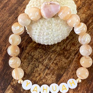 May include: A beaded bracelet with the word "YAHUAH" spelled out in gold letters on white beads. The bracelet features peach-colored, round beads and a larger pink stone. The bracelet is displayed on a textured, off-white surface.