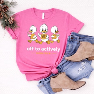 Op de afbeelding: Een roze t-shirt met drie cartoon eendjes die rennen. De eenden dragen oranje shirts en houden verschillende voorwerpen vast. De tekst "off to actively" en "hustle" staat op het shirt. Het shirt wordt gecombineerd met een blauwe jeans en beige laarzen.