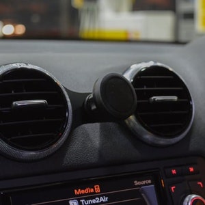 Puede incluir: Un interior de coche negro con dos salidas de aire y un soporte magnético para teléfono. La pantalla de la radio del coche muestra "Media 6" y "Tune2Air".