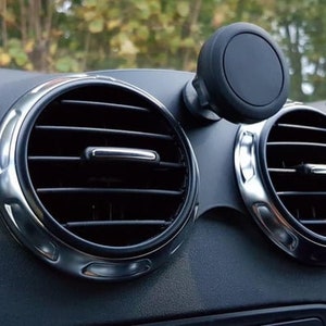 Puede incluir: Un soporte magnético negro para teléfono móvil fijado a la ventilación del salpicadero de un coche. El soporte está diseñado para sujetar un teléfono inteligente de forma segura mientras se conduce.