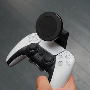 Peut inclure: Une manette de jeu PlayStation 5 DualSense blanche et noire avec un support de téléphone magnétique noir fixé sur le dessus.