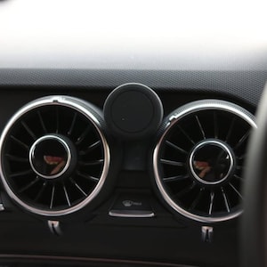 May include: Close-up of a car dashboard featuring two circular air vents with silver trim and a black center. A black magnetic phone mount is positioned between the vents. The dashboard is black with a textured surface, and the steering wheel is visible on the right.