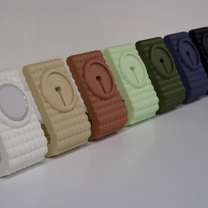 May include: A set of seven phone stands in various colors, including white, beige, brown, green, light green, blue, and black. Each stand has a raised circular area for wireless charging.