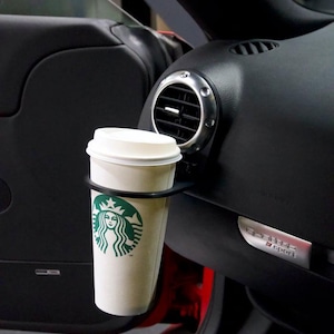 Może przedstawiać: Czarny uchwyt na kubek samochodowy z białym kubkiem kawy Starbucks w środku. Uchwyt na kubek jest przymocowany do boku deski rozdzielczej samochodu. Wnętrze samochodu jest czarne i czerwone.