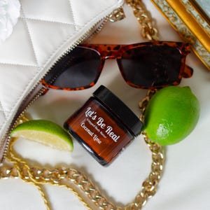 May include: An overhead shot features a white quilted handbag with a zipper, tortoise shell sunglasses, a jar of "Let's Be Real" Coconut Lime whipped body butter, a gold chain, a lime wedge, a whole lime, and a gold framed mirror.