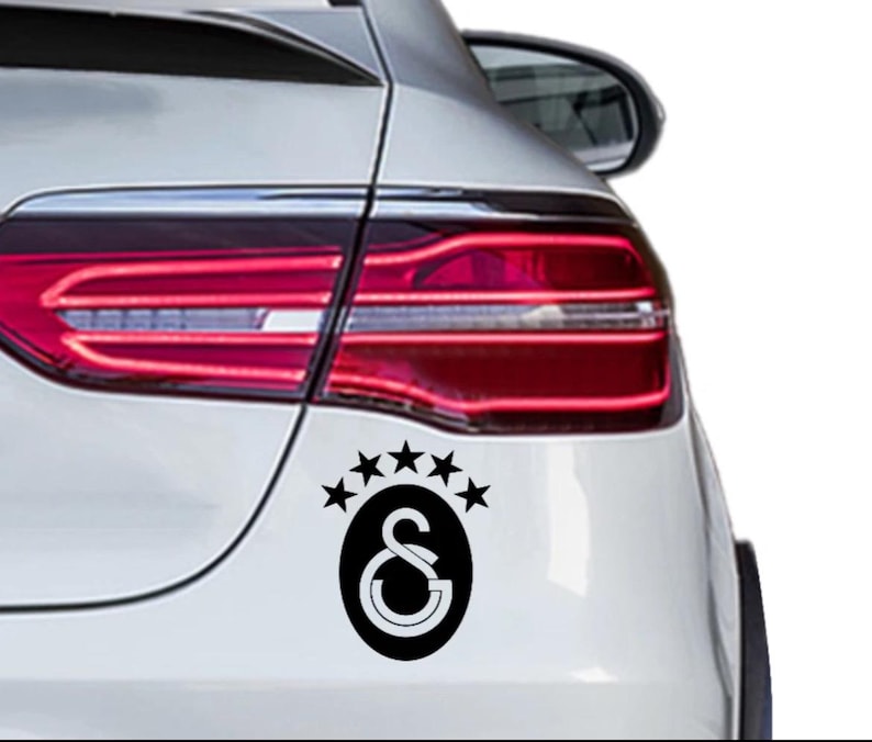 May include: A black and white decal featuring the Galatasaray logo, a stylized "S" and "G" inside an oval, with five stars above. The decal is affixed to the rear of a white car, near the taillight.