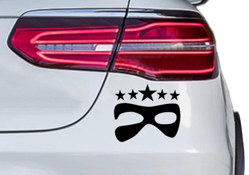 Peut inclure: Autocollant de voiture en vinyle noir repr&eacute;sentant un masque stylis&eacute; surmont&eacute; de cinq &eacute;toiles. L'autocollant est appliqu&eacute; &agrave; l'arri&egrave;re d'une voiture blanche, pr&egrave;s du feu arri&egrave;re.