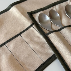 May include: A beige utensil roll with dark green trim and stitching, holding several stainless steel spoons. The roll is made of a soft, felt-like material and is partially open, displaying the spoons.