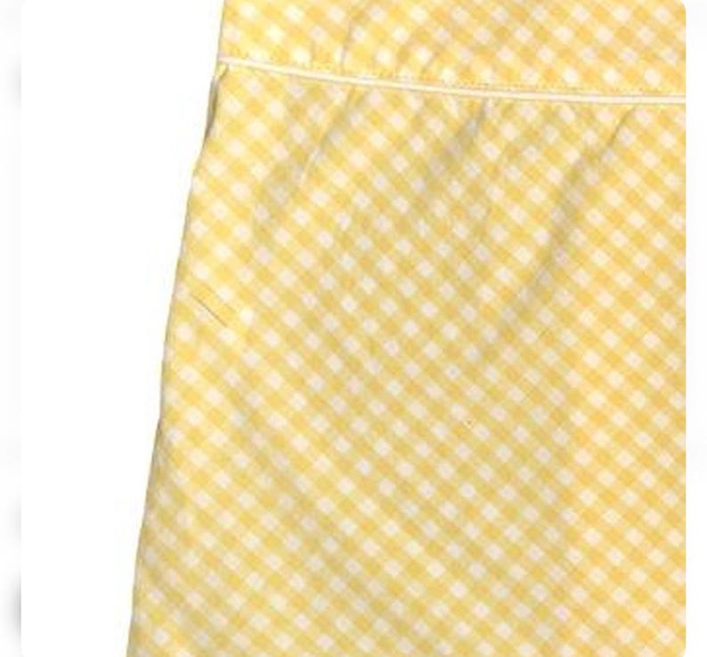 May include: Close-up of a yellow and white gingham fabric. The fabric is a classic checkered pattern, with a small white border at the top. The fabric is likely cotton or a similar material, and is suitable for clothing or home decor.
