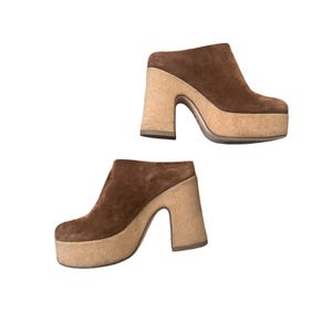 May include: Two brown suede platform clogs with cork heels. The clogs have a rounded toe and a slip-on design. The heels are approximately 4 inches high.