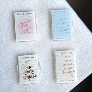 以下が含まれることがあります： 図書館をテーマにした4枚の透明アクリルカード。2枚のカードには「Guest Check」と「buy me books」や「I'm pretty」などのフレーズが書かれています。他の2枚は「Library Card」と「one more chapter」と「The Tortured Readers Department」と書かれています。