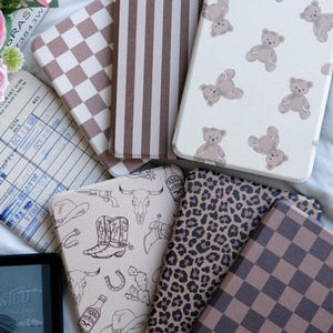 Könnte beinhalten: Eine Sammlung von sechs rechteckigen E-Reader-Hüllen mit verschiedenen Mustern, darunter braun-weißes Schachbrettmuster, vertikale braune Streifen, ein Teddybären-Druck, ein Western-Motiv mit Stiefeln und Schädeln sowie ein Leopardenmuster.