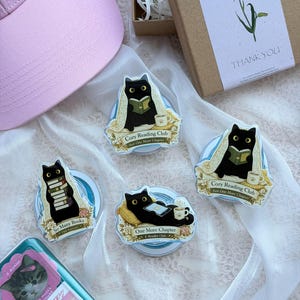 Puede incluir: Tres pegatinas circulares con gatos negros leyendo libros o lectores electrónicos con el texto Cozy Reading Club, So Many Books y One More Chapter. Las pegatinas están dispuestas cerca de una gorra rosa y una caja de regalo marrón con flores.