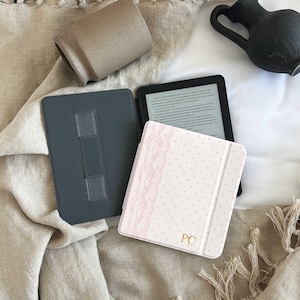 May include: An e-reader device in a light pink case with a white polka dot pattern and a vertical strip of pink lace trim. The case features the gold-colored initials PC on the front cover. The device is open, revealing a dark gray interior.