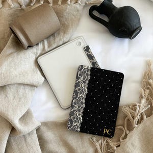 Puede incluir: Una funda para lector electrónico negra con un borde de encaje blanco en el lado izquierdo y un patrón de puntos blancos sobre la tela negra, con las iniciales doradas PC impresas en la esquina inferior derecha, junto a una funda blanca lisa.