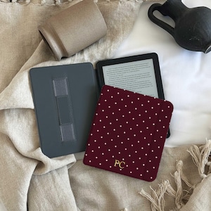 May include: An e-reader with a burgundy cover featuring white polka dots and gold initials, next to a gray case and a beige ceramic cup. The items are arranged on a beige fringed textile.