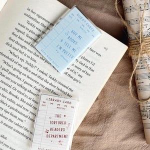 Może przedstawiać: Dwie ozdobne karty z tekstem, jedna jasnoniebieska z napisem "Buy Me Books & Tell Me I'm Pretty", a druga biała z napisem "The Tortured Readers Department". Karty leżą na otwartej książce.