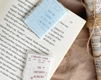 May include: Two rectangular paper cards resting on an open book. The top card is light blue with the text GUEST CHECK, BUY ME BOOKS & TELL ME I'M PRETTY, and READ MORE BOOKS. The bottom card is white with the text LIBRARY CARD, THE TORTURED READERS DEPARTMENT, and READ MORE BOOKS.
