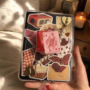 Puede incluir: Una funda protectora transparente para tableta o lector electrónico decorada con varias pegatinas, incluyendo un reloj digital rojo, un pastel de cerezas, un patrón de cuadros rojos y blancos, una mariposa, una cesta y un sobre con el texto P.S. I love you.