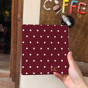 May include: A burgundy tablet case with a white polka dot pattern. The case has a gold-colored "LC" monogram in the lower right corner. The case is held in a person's hand.
