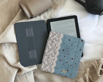 Puede incluir: Una funda para lector electrónico azul con un borde de encaje blanco con patrones de lazos en el lado izquierdo y pequeñas estrellas de color rojo oscuro. La funda incluye las letras doradas del monograma PC y se muestra abierta con interior gris.