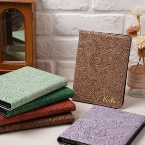 May include: A collection of rectangular passport holders featuring an embossed floral pattern. The covers come in light green, dark green, red, brown, and purple. One brown holder displays the gold-embossed initials K.K. on the bottom right corner.