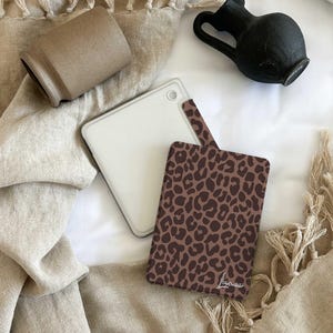 以下が含まれることがあります： 白いタブレットケースと、ハンドル付きの茶色のヒョウ柄タブレットケースの俯瞰図。小さな茶色の花瓶と黒い水差しも表示されています。ケースはベージュ色の表面にあります。