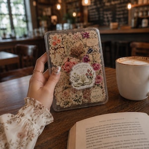 Op de afbeelding: Een doorzichtig rechthoekig plastic doosje gevuld met diverse bloemen- en plantenstickers. Een centrale ronde sticker toont een illustratie van een open boek met de tekst "BOOKS ARE MY HAPPY PLACE" en "ROMANCE" op een klein label.