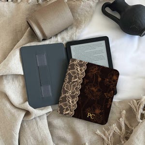 Puede incluir: Un lector electrónico con una funda de color marrón oscuro con un patrón de flores y mariposas, acentuado por un borde beige similar al encaje en el lado izquierdo y las iniciales PC en color dorado impresas en la esquina inferior derecha.