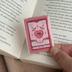 May include: A rectangular acrylic pin featuring a pink tarot card design titled The Lovers. The illustration depicts a central heart with a single eye, pierced by two swords, surrounded by clouds, stars, and decorative borders in shades of pink.