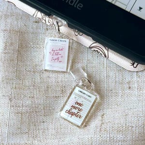 May include: Two clear acrylic charms with silver key rings. One charm reads "Guest Check" with text "buy me books, tell me I'm pretty." The other charm reads "Library Card" with text "one more chapter."