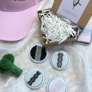 May include: A light pink baseball cap with the text "ls read" in red embroidery, a brown cardboard gift box with white paper filler, and four circular pins featuring black, brown, and pink lace patterns on polka dot backgrounds.