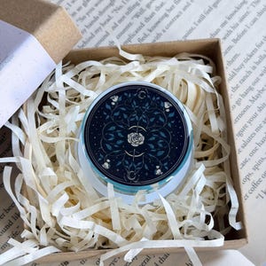 May include: A circular glass paperweight featuring a dark blue background with a white rose illustration, intricate vine patterns, and small crescent moon symbols, nestled inside a square cardboard gift box filled with white paper shred.