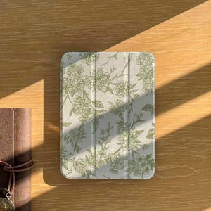 May include: A light-colored tablet case with a floral pattern in shades of green. The case is rectangular and has a folding cover. A brown leather-bound journal with a decorative charm is visible on the left.