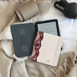 May include: A cream-colored e-reader case with black polka dots, featuring a dark red lace trim on the left side and the gold-colored initials PC embossed on the bottom right corner. The case is open, revealing a gray interior with a hand strap.