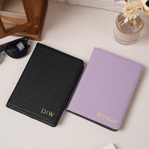 May include: Two rectangular passport holders with a textured finish. One holder is black with the gold embossed text D|W, and the other is light purple with the gold embossed name Krystian. Both items feature stitched edges and a slim profile.