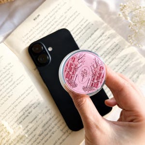 May include: A circular phone grip featuring a pink background with a dark red line art illustration of city buildings, bare trees, and a central bouquet of flowers. The accessory is attached to a black smartphone resting on an open book.