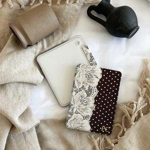 May include: A rectangular passport holder featuring a dark brown fabric with white polka dots on one side and a white floral lace overlay on the other. The bottom right corner displays the gold embroidered initials PC on the dark brown fabric.