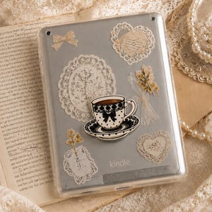 Op de afbeelding: Een zilveren Kindle e-reader in een doorzichtige beschermhoes versierd met diverse stickers, waaronder een zwart-witte theekop, kanten hartjes, een klein strikje en botanische illustraties. Het woord Kindle staat onderaan op het apparaat.