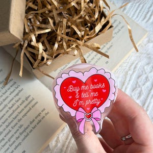 Könnte beinhalten: Ein herzförmiger Acryl-Anhänger mit roter Mitte und dem weißen Text "Buy me books & tell me I'm pretty", umgeben von einem rosa gewellten Rand, kleinen roten Herzen und einer rosa Schleife an der Unterseite, gehalten vor einem Buch und einer Geschenkbox.