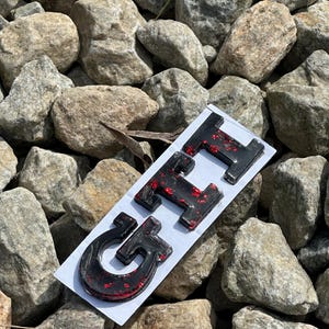 Peut inclure: Emblème GT noir et rouge sur une carte blanche, sur fond de roches grises. Les lettres sont brillantes, avec des accents de paillettes rouges. L'emblème est probablement pour une voiture ou un véhicule.