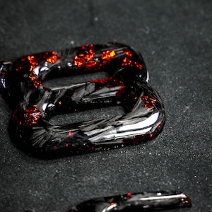 May include: Close-up of glossy black and red decorative letter shapes. The letters have a textured, carbon fiber appearance with embedded red glitter, creating a striking contrast against the dark background. The letters are likely part of a larger set.