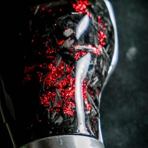 Puede incluir: Primer plano de un pomo de palanca de cambios negro y rojo. El pomo tiene un aspecto de fibra de carbono con acentos rojos brillantes. Una banda plateada rodea la parte superior de la base cosida en cuero. El fondo es oscuro.