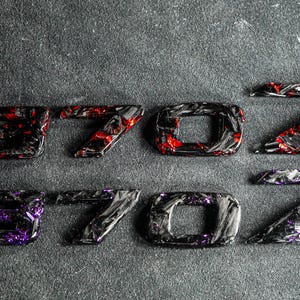 Peut inclure: Deux ensembles d'emblèmes de voiture épelant "370Z". Un ensemble a des accents rouges, l'autre a des accents violets. Les emblèmes sont noirs avec un aspect fibre de carbone, sur un fond gris foncé texturé.