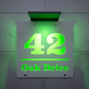 以下が含まれることがあります： 明るい緑色の「42」と「Oak Drive」の文字がグレーの背景に表示された、照明付きの住所看板。看板は壁に取り付けられており、上部にソーラーパネルが付いています。