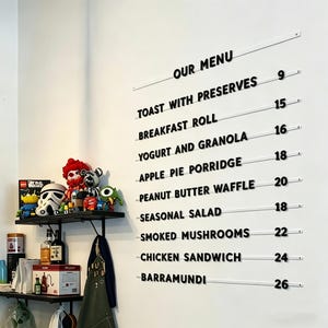 May include: A white wall displays a black and white menu with items like "Toast with Preserves" and "Chicken Sandwich." A shelf below holds toys and bottles. The menu is in a restaurant or cafe.