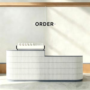 Puede incluir: Un mostrador de recepción moderno blanco y gris con un diseño curvo. El mostrador tiene un patrón texturizado de líneas verticales y una encimera con tazas y vasos. La palabra "ORDER" se muestra encima del mostrador.