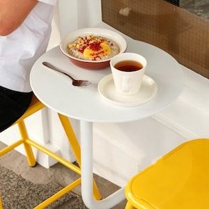 May include: A white, round table with a bowl of oatmeal topped with fruit and nuts, a cup of tea, and a fork. The table is supported by a white leg and is next to a yellow chair. The scene suggests a cafe or breakfast setting.