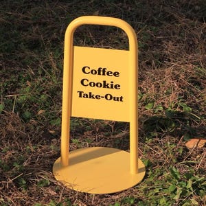 Puede incluir: Un letrero amarillo con las palabras "Coffee Cookie Take-Out" en negro. El letrero tiene una parte superior redondeada y una base circular. Está colocado sobre césped, indicando un servicio de comida para llevar.