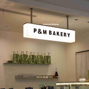 May include: A white rectangular sign with black lettering that reads "P&M BAKERY" hanging from the ceiling. The sign is illuminated with white LED lights.